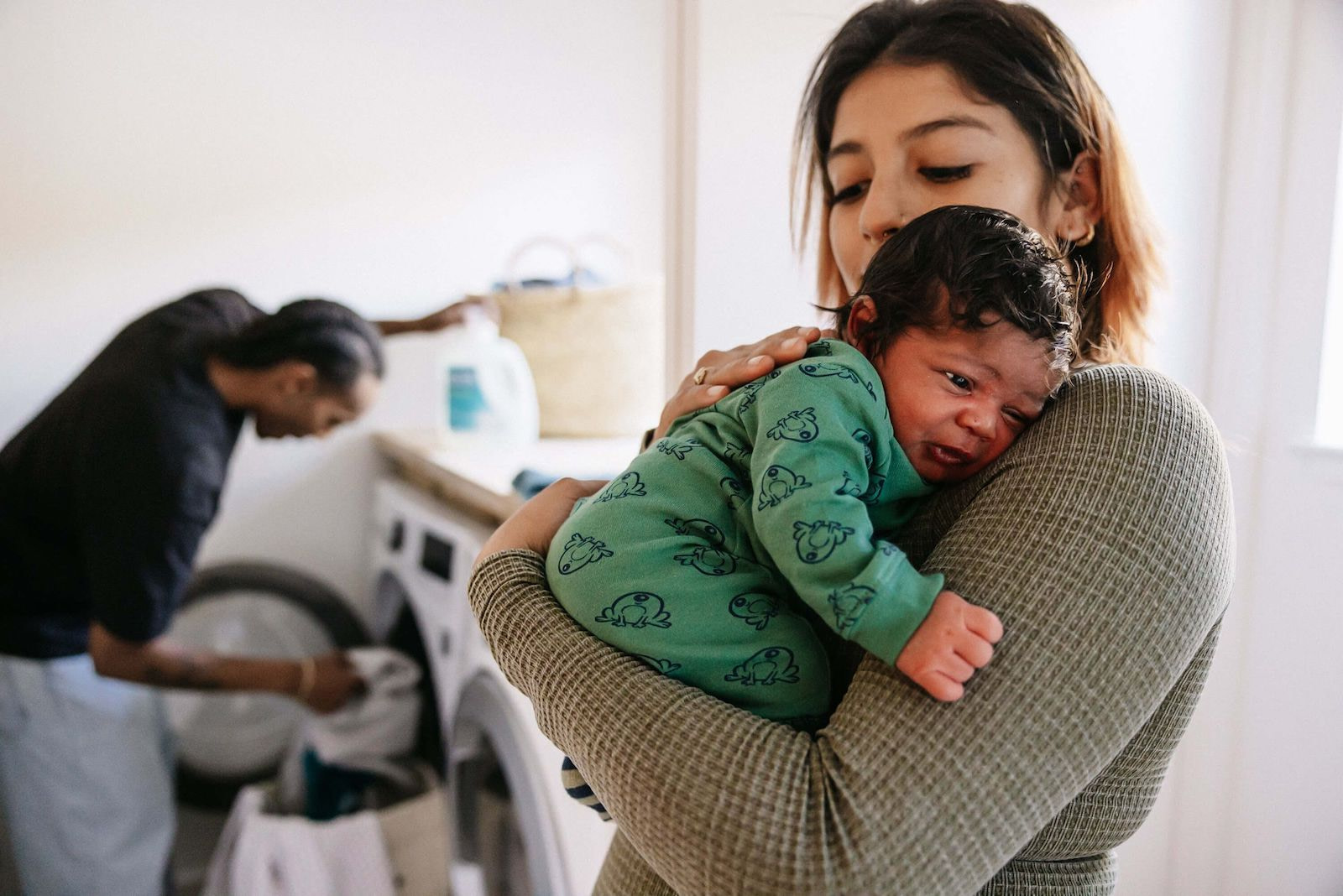 Mother Holding Baby with Partner Doing Laundry
